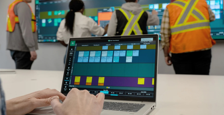 Hands operate a laptop displaying a colorful industrial control dashboard as workers in high-visibility vests monitor screens in a control room.