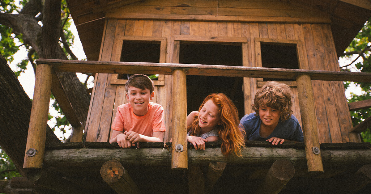 Three children smiling while peering down from a wooden tree fort.