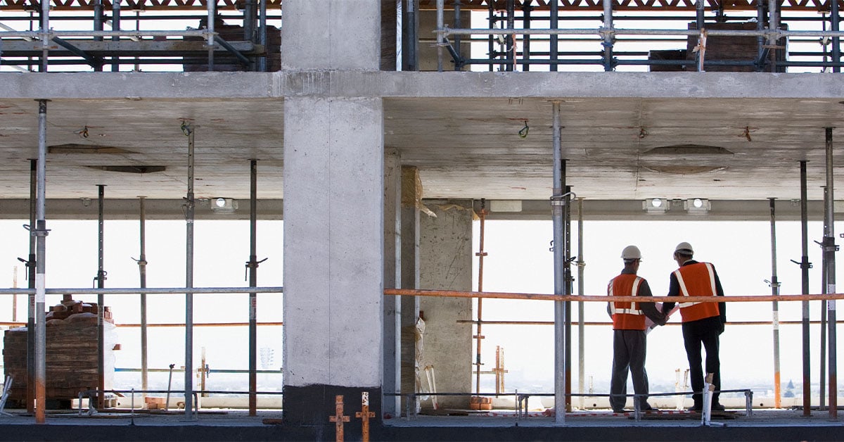 Two construction workers wearing hard hats and hi-vis vests assessing progress on a construction site.