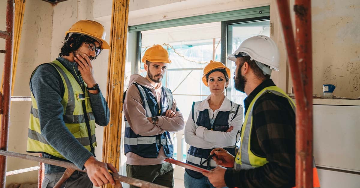 Four construction workers wearing hard hats and hi-vis vests engage in a discussion on a jobsite, with one of them holding a clipboard and pen.