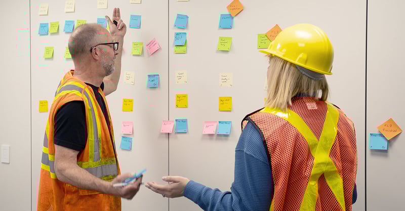 Two tradespeople stand in front of a sticky-note wall keeping track of a Lean project